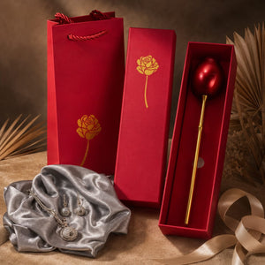 A red gift set containing a silver-toned pendant necklace draped on a satin cloth, a tall red gift bag with a gold rose print, a matching red box, and an open red presentation box holding a gold-toned rod with a red spherical top, styled with dried palm leaves and ribbon on a brown surface.