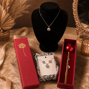 A jewelry display set on a beige fabric surface, featuring a silver pendant necklace on a black velvet bust, a red gift box with a gold rose design, a pair of silver earrings on a white card, and a red gift box containing a red apple-shaped pen or accessory.